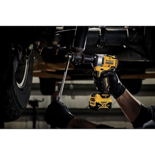 A person wearing black gloves using a yellow and black DEWALT DCF901P1 cordless impact wrench to work on a vehicle undercarriage in a workshop setting.