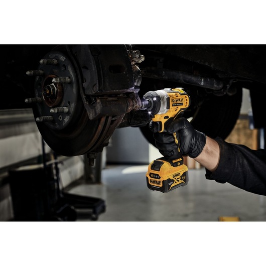 A person wearing a black glove uses a DEWALT cordless impact wrench to work on a vehicle's wheel hub assembly in a garage.