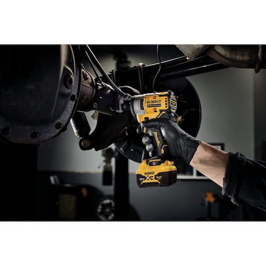 A person wearing black gloves uses a DEWALT DCF901P1 12V XR cordless impact wrench to work on a vehicle's axle in a garage setting.