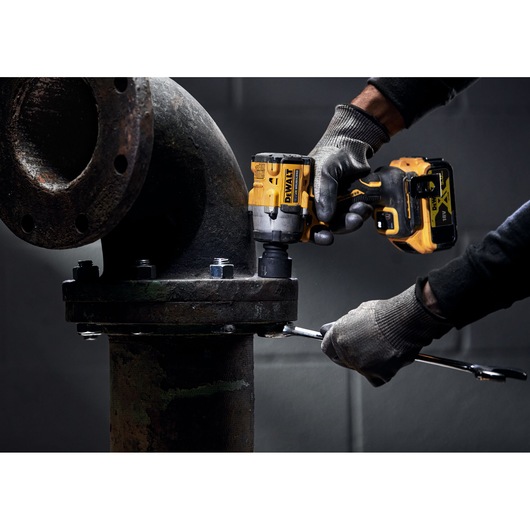 A person wearing gloves uses a DEWALT cordless impact wrench to tighten bolts on a large industrial pipe.
