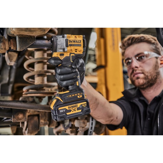 A person wearing black gloves is using a DEWALT 18V XR cordless impact wrench to work on a vehicle's suspension system in a workshop. The person's face is blurred.