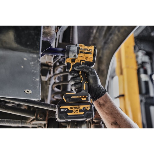 A person wearing a black glove is using a DEWALT 18V XR cordless impact wrench to work on the underside of a vehicle.