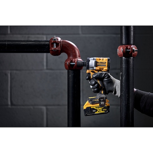 A person wearing black gloves uses a DEWALT cordless impact wrench to tighten a bolt on a metal pipe joint. The tool is yellow and black, and the battery pack is visible. The background features a grey brick wall and industrial piping.