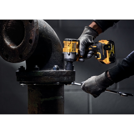 A person wearing gloves uses a DEWALT cordless impact wrench to tighten bolts on a large industrial pipe flange.
