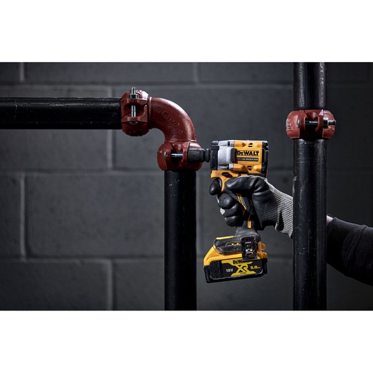 A person wearing gloves uses a DEWALT cordless impact wrench to fasten a bolt on a metal pipe fitting. The tool is yellow and black, and the scene is set against a dark, industrial background.