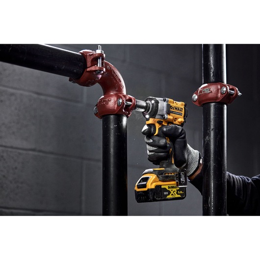 A person wearing work gloves uses a yellow and black DEWALT cordless impact wrench to tighten a bolt on a black metal pipe with a red fitting.