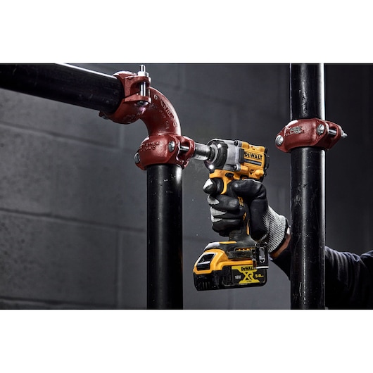 A person using a yellow and black DEWALT cordless impact wrench to work on a metal pipe joint, wearing black gloves. The tool is being used to tighten or loosen bolts in an industrial setting with black pipes and red pipe fittings.