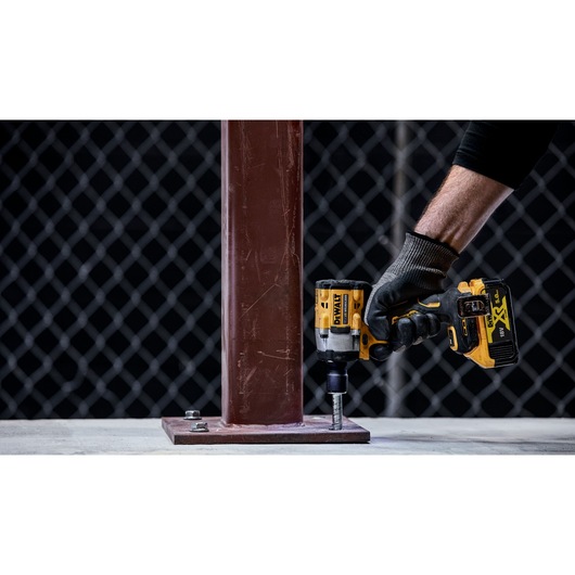 A person wearing a glove uses a DEWALT cordless impact wrench to tighten a bolt securing a metal beam to a concrete surface.