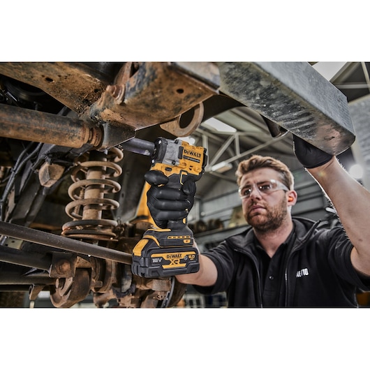 A person wearing black gloves uses a yellow and black DEWALT DCF923N cordless impact wrench to work on a metal automotive suspension part in a workshop. The person’s face is blurred.