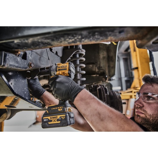 A person wearing black gloves uses a DEWALT DCF923P2G 18V cordless impact wrench to work on the suspension components under a vehicle in a workshop.