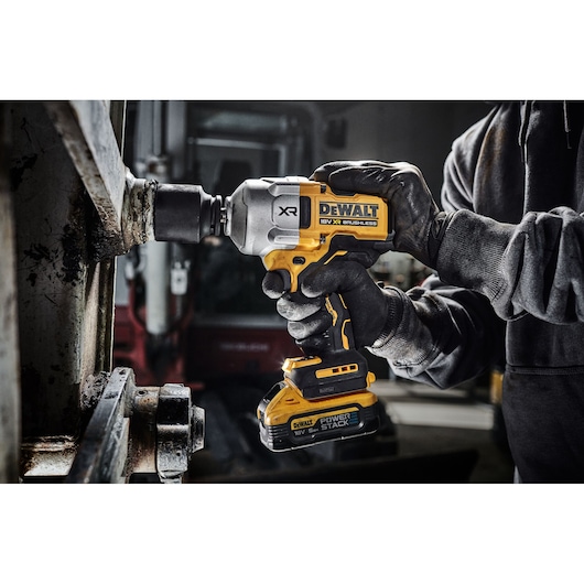 A person wearing black gloves is using a DEWALT 18V XR Brushless impact driver with POWERSTACK battery to work on a metal surface in an industrial setting.