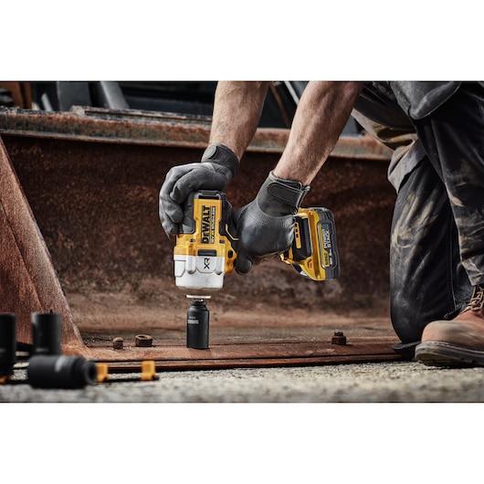 A person wearing work gloves uses a yellow and black DEWALT cordless impact wrench to loosen a bolt on a rusty metal surface at an industrial site.