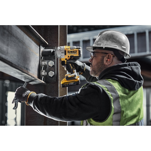 A person wearing a safety helmet and high-visibility vest is using a DEWALT DCF961H2T cordless impact wrench to tighten bolts on a steel structure at a construction site.