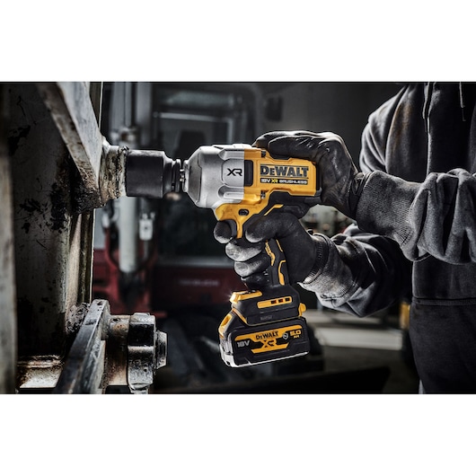 A person wearing gloves uses a DEWALT DCF961N 18V XR brushless impact driver to tighten or loosen a large bolt on a metal structure.