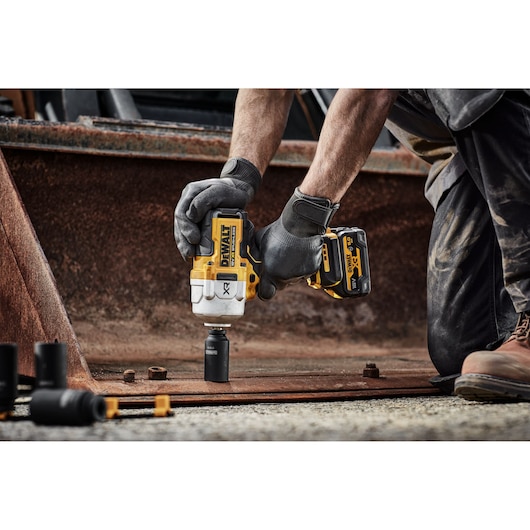 A person wearing work gloves uses a DEWALT cordless impact wrench to fasten or loosen a bolt on a metal plate at a construction site. The tool features yellow and black DEWALT branding.