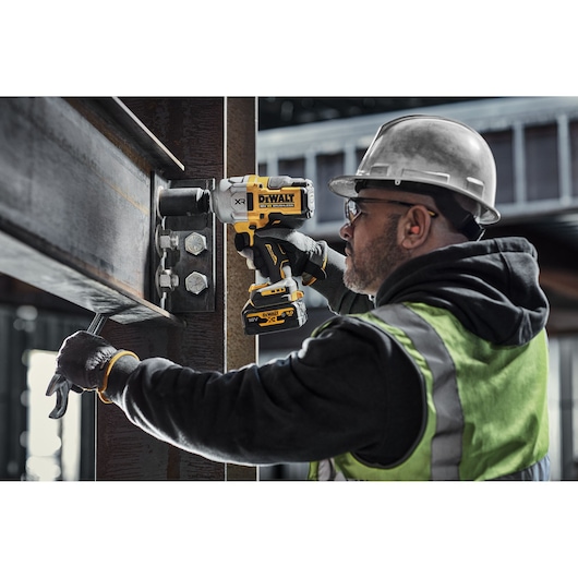 A person wearing protective gear uses a DEWALT DCF961N cordless power tool to fasten bolts on a large steel beam at a construction site.