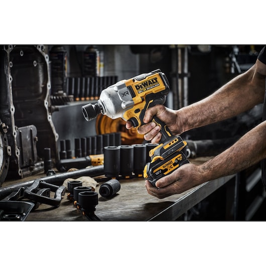A person holds a DEWALT cordless impact wrench in a workshop setting, surrounded by various tools and accessories on a workbench.