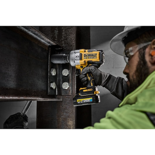 A person in work attire uses a DEWALT DCF964NT cordless impact wrench to tighten large bolts on a steel beam at an industrial construction site. The person's face is not clearly visible.