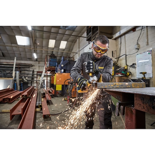 A person using a DEWALT portable grinder to cut metal, with sparks flying in an industrial workshop setting. The person's face is blurred for privacy.