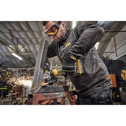 A person wearing protective gloves uses a DEWALT DCG404NT cordless angle grinder to cut metal, producing sparks, in an industrial workshop setting. The person's face is obscured.