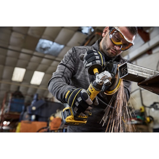 A person wearing safety gloves uses a DEWALT cordless angle grinder on a metal pipe, producing sparks in an industrial workshop setting. The person's face is blurred.