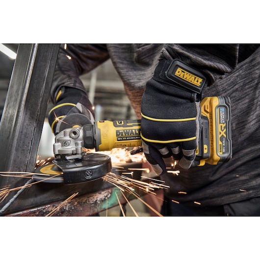 A close-up of a person wearing gloves and operating a DEWALT cordless angle grinder, creating sparks while grinding a metal surface.