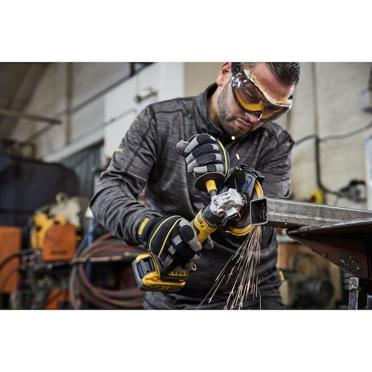 A person wearing protective gloves uses a DEWALT cordless angle grinder (DCG404NT) to cut a metal beam in a workshop. Sparks are flying from the metal, indicating active use of the tool. The person's face is blurred.