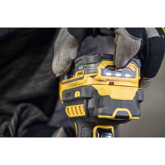 Close-up of a gloved hand pressing a button on a DEWALT cordless power tool. The LED indicator lights on the tool are illuminated.