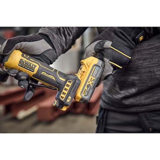 Close-up of a person wearing protective gloves holding a yellow DEWALT cordless tool, specifically an angle grinder with a brushless motor and XR battery, in an industrial setting.