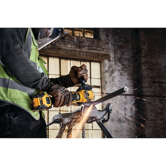 A DEWALT cordless grinder cutting metal, sparks flying, worker wears gloves and safety gear in industrial setting.
