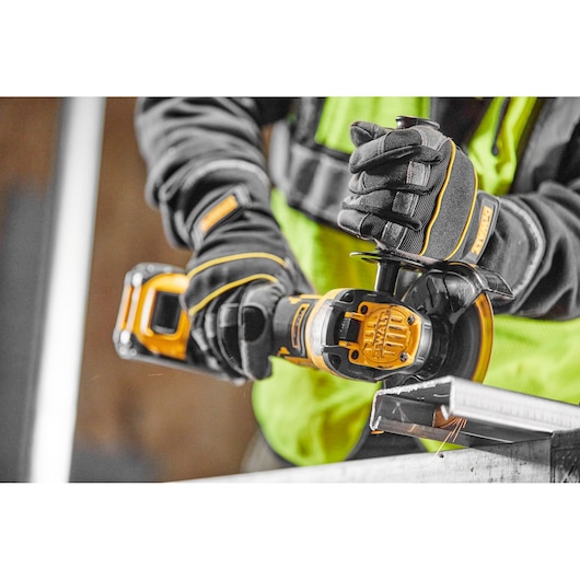 Gloved hands using a DEWALT cordless grinder on a metal piece, worker wearing a safety vest.