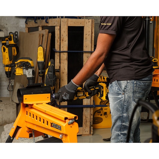 A person wearing gloves and a black shirt is operating a DEWALT power tool, working on a piece of metal secured in a yellow workbench. Other DEWALT tools and construction materials are visible in the background.