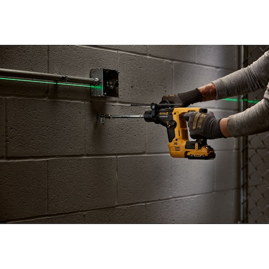 A person using a yellow DEWALT cordless rotary hammer drill to install electrical conduit on a concrete block wall. The user is wearing gloves and aligning the conduit with a visible green laser line.