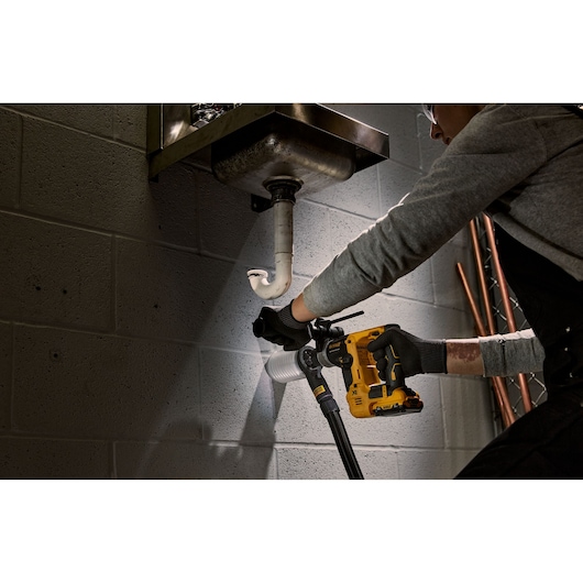 A person wearing gloves uses a DEWALT cordless rotary hammer drill (DCH072NT) to drill into a wall near plumbing pipes under a metal sink.