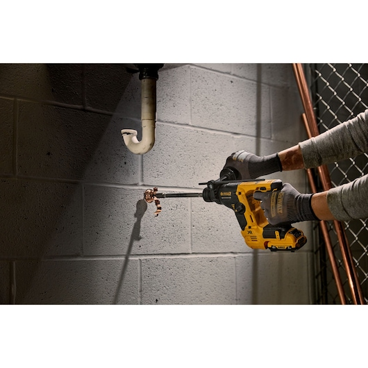 A person wearing gloves uses a yellow DEWALT rotary hammer drill (model DCH072NT) to drill into a concrete wall near a plumbing pipe.