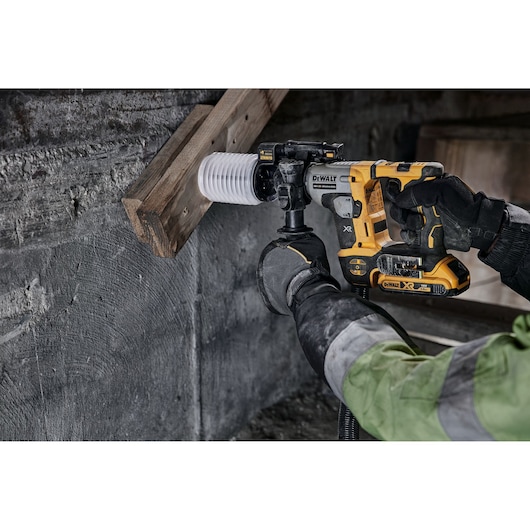 A person wearing gloves and a work jacket uses a DEWALT cordless rotary hammer drill to drill into a concrete wall, with a dust extraction attachment connected to the tool.