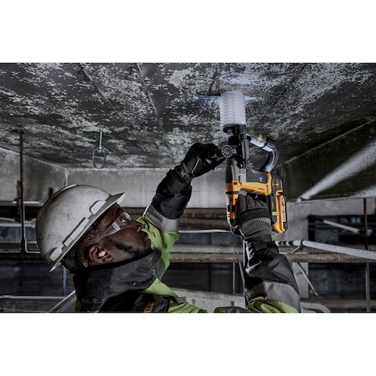 Person wearing safety gear uses a DEWALT cordless rotary hammer drill to drill into a concrete ceiling in an industrial setting.