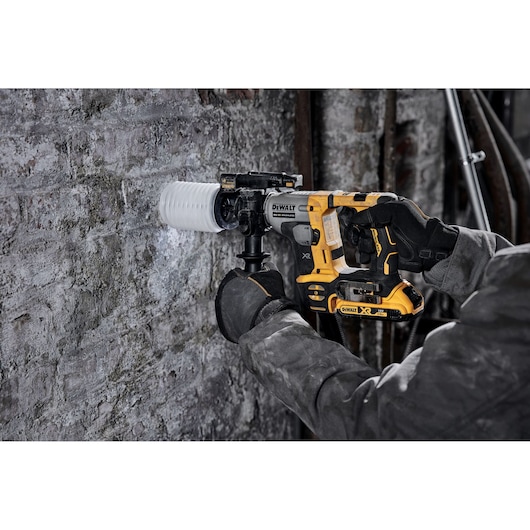 A person wearing work gloves operates a DEWALT DCH172N cordless rotary hammer drill with a dust extraction attachment, drilling into a concrete wall.