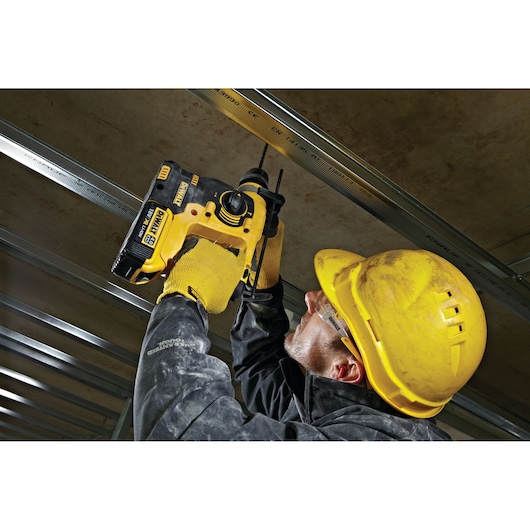 Worker drills overhead using a DEWALT DCH253N rotary hammer drill, wearing yellow gloves and a yellow safety helmet.