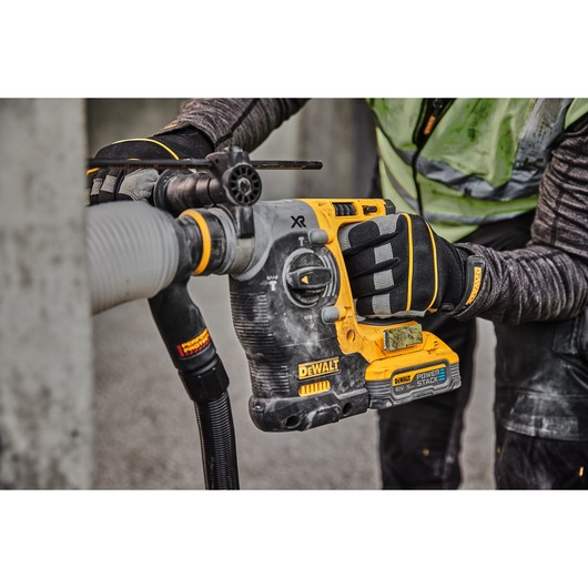 A close-up of a worker using a DEWALT cordless rotary hammer drill with a POWERSTACK battery to drill into concrete, wearing protective gloves and clothing.