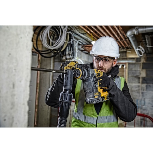 Worker uses a DEWALT rotary hammer drill on a construction site wearing safety gear and a high-visibility vest.