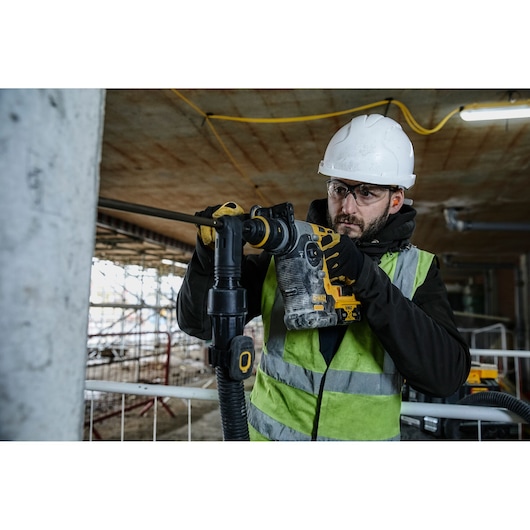 Worker using DEWALT DCH274N tool to drill into concrete at a construction site, wearing safety gear and vest.