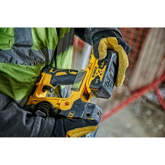 Gloved hand holds a DEWALT cordless power tool with an 18V XR lithium battery at a construction site.
