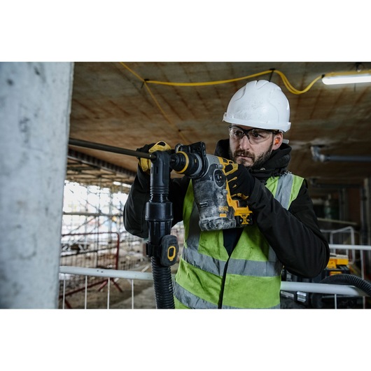 Worker using a DEWALT rotary hammer drill on a construction site, wearing a helmet, gloves, and safety vest.
