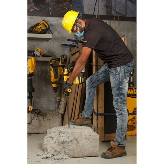 A person wearing a yellow safety helmet, gloves, and work boots is using a DEWALT DCH333 rotary hammer drill to work on a large concrete block indoors. The setting includes additional construction tools and materials visible in the background.