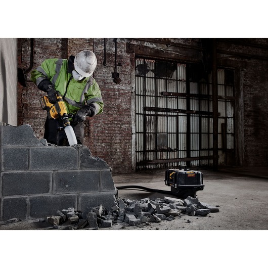 Worker uses DEWALT power tool to demolish concrete wall in an industrial setting with rubble on the ground.