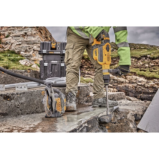 Person using a yellow DEWALT rotary hammer drill to break concrete outdoors, with additional DEWALT tools and toolboxes visible in the background.