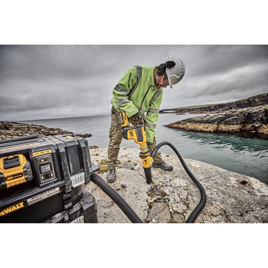 A construction worker wearing a green jacket, safety helmet, and ear protection is using a yellow DEWALT rotary hammer on a rocky surface near a body of water. A DEWALT equipment box is visible in the foreground with a hose connected to the tool.