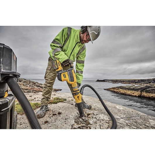 A person wearing a green safety jacket, gloves, and a helmet is using a yellow DEWALT rotary hammer drill to break concrete outdoors near a body of water. A vacuum hose is attached to the tool to collect dust.