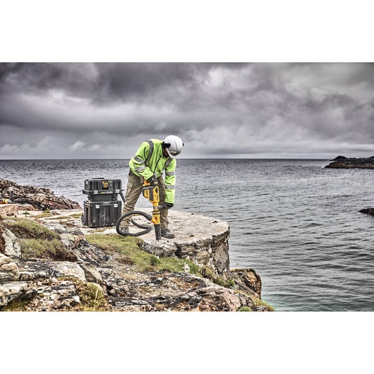 A worker in a high-visibility jacket and helmet is using a yellow DEWALT demolition tool on a rocky cliff above the ocean, with DEWALT storage boxes nearby. The sky is cloudy.